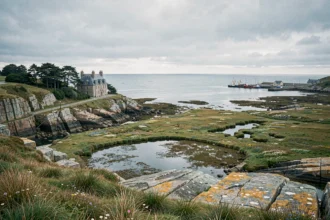 Paysage côtier breton avec falaises de granit, manoir en pierre et baie atlantique calme sous un ciel nuageux